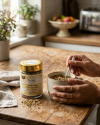 Person adding herbs to a mug on a wooden table with a jar of chamomile tea in the background.