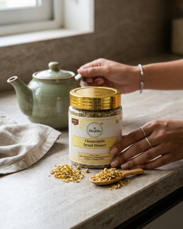 Person holding a jar of chamomile dried flowers on a kitchen counter with a teapot in the background.