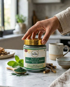 Person holding a jar of Deshla Spirulina powder on a kitchen counter with ingredients around.