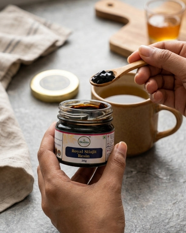 Person holding a jar of 'Royal Silajit Resin' with a spoonful above a cup of tea on a textured surface.
