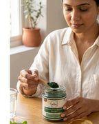 Woman using a spoon to scoop green powder from a jar labeled 'Desha' on a wooden table.